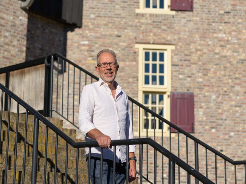 Man van middelbare leeftijd met bril poseert op trappen monumentaal pand. Hij draagt een witte blouse.
