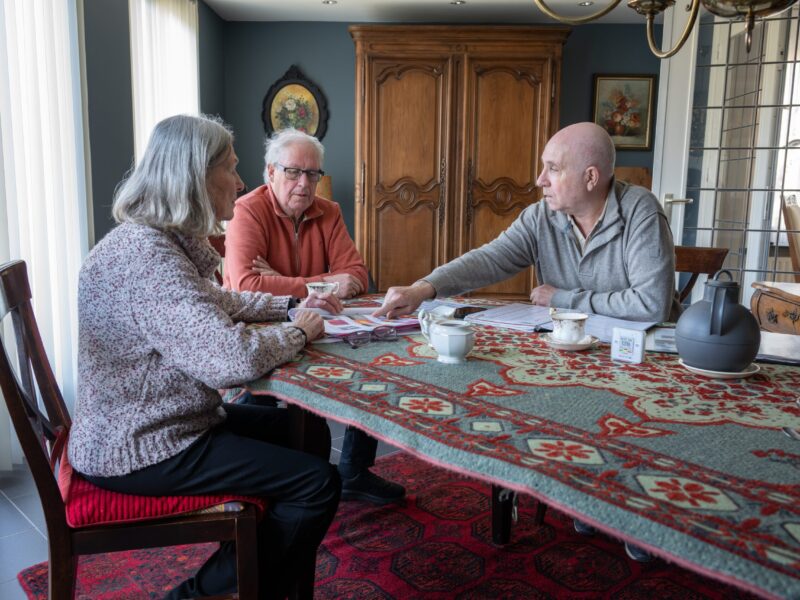 Drie mensen zitten aan een tafel. Ze praten over informatie uit een folder die op de tafel ligt.